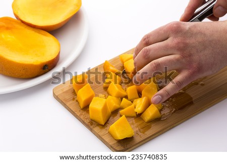 Yellow fruit flesh of a ripe mango being cut into dice on a wooden cutting board. Finger tips of a left hand fixing the juicy pulp pieces, while a right hand is pulling a knife. White background.