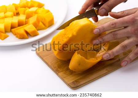 Middle third of a mango containing its fruit pit is being separated from its skin. Left finger tips bedewed with mango juice are balancing the pulp, while a right hand is moving a knife. White ground.