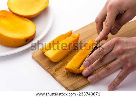 One of the two outer thirds of a mango divided into three slices is being cut into four pieces. One hand is positioning the fruit chips, while the other is guiding a kitchen knife. White background.