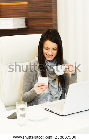 Businesswoman drinking coffee at desk, looking at mobile phone, smiling in the office