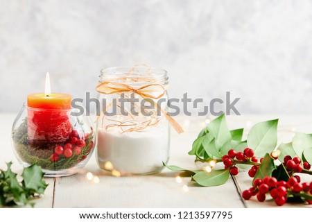 Christmas Table Decoration. Fairy lights in a jar next to vase with holly berries and red candle.