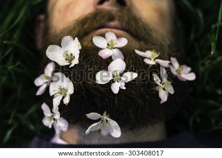mans head laying in the grass with flowers tucked into his beard with a shallow depth of field with a instagram filter