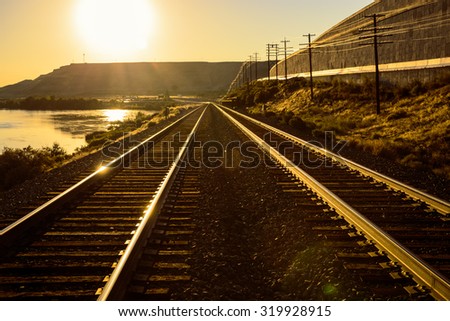 Pair of train tracks laid along the snack river passing through glenns ferry during sunrise or sunset with a sun flare and mountains in the background