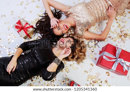 Two elegant  gorgeous women in trendy sequin dress lying on white floor with shining golden confetti and red gift boxes . Celebrating new year or birthday party.