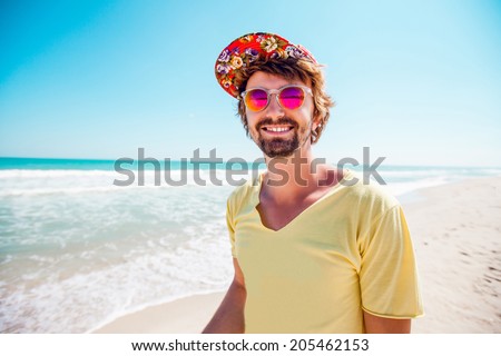 Close portrait stylish man with glasses and colored cap smiling and relaxing on a tropical beach. Sunny summer colors.
