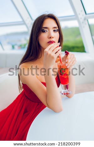 Beautiful sexy  lady  sitting alone  in a restaurant in an elegant red dress and drinking a cocktail overlooking the city.