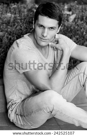 Handsome and attraktive man posing outdoor . Sitting on the ground and looking at camera . Black and white colors.