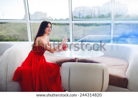 Beautiful glamour  woman  sitting alone in a restaurant in an elegant red dress and drinking a cocktail overlooking the city.