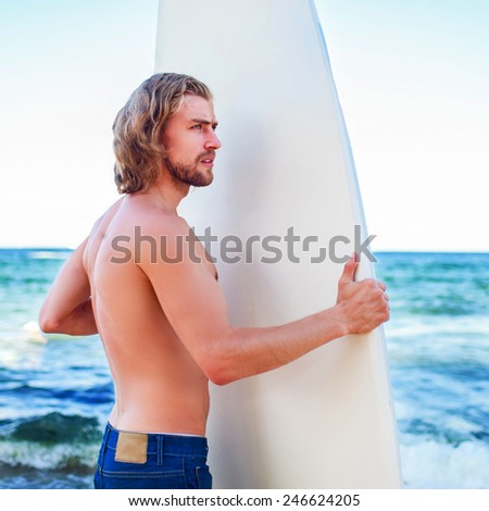 Young attractive surfer sports man  with long hair wearing jeans shorts   and looking at the horizon while carrying his surfing board during a sunny day against the intense blue sky.