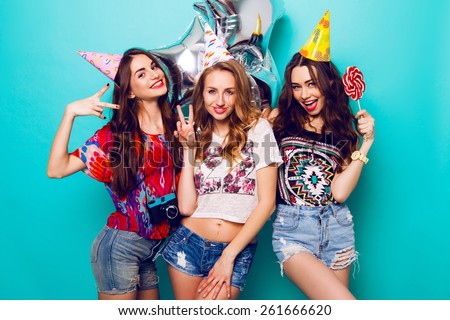 Three beautiful  happy sexy woman  in  stylish summer outfit , paper hats and purity balloons having fun and celebrate  birthday.  Colorful blue background . Pretty girl  hold big lollypop.