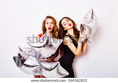Two  funny friends with red lips in evening black and red dress on the party  make  grimace and have fun .  Two  pretty emotional girls hold balloons and posing against white wall.