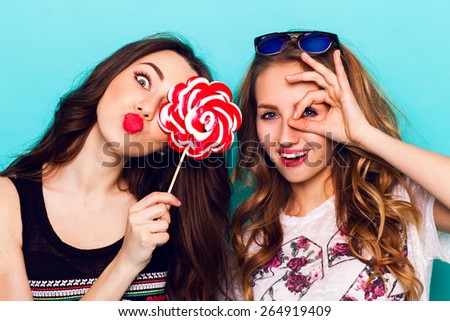 Close op fashion portrait of Two friends have fun , make grimaces ,fun emotions and shows the hand signs.  Pretty girls wearing summer clothes and  posing against blue wall.