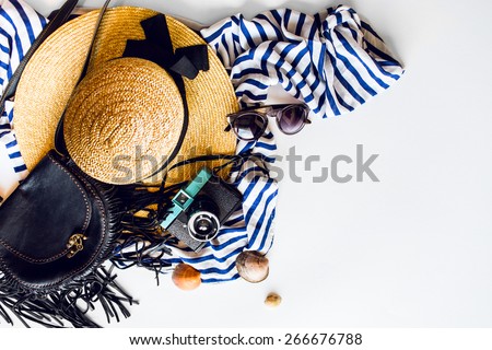 Top view of summer accessories for modern woman on her vacation. Straw hat,  camera, stylish sunglasses, black leather boho bag and striped beach dress on white floor.