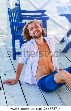 Natural outdoor  fashion portrait of great looking young man with big smile  sitting on the wooden floor on  beach caffe. Warm sunshine colors.