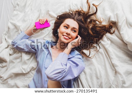 Over head portrait view of an beautiful  young woman laying down on her bed in her home bedroom using an listen to music on phone  with her headphones. Have curly hair, perfect smile and enjoying .