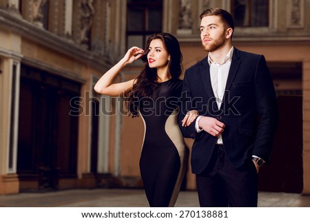 Couple in love in elegant outfit walking in old town street . Pretty brunette woman with red lips and her handsome boyfriend have time off in the evening.