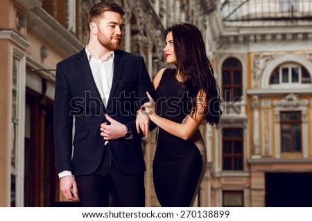 Attractive couple in love in elegant glamour  outfit walking in old town street . Pretty brunette woman with red lips  in long cocktail  dress and her handsome boyfriend have time off.