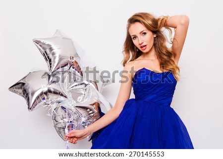 Pretty young women celebrate  birthday  and holding  silver party balloons. Wearing blue evening stylish  dress and have day of against white wall.
