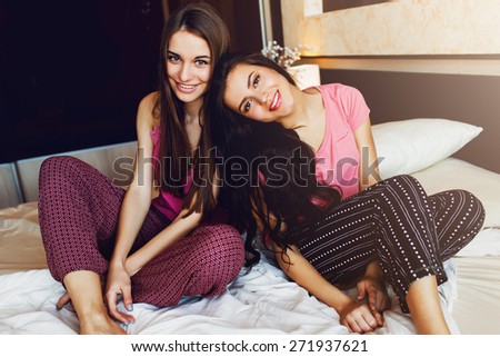 Close up lifestyle indoor fresh  portrait of two pretty smiling  friends . Two girls sitting in bed wearing  pink pajamas and enjoy their  good time.