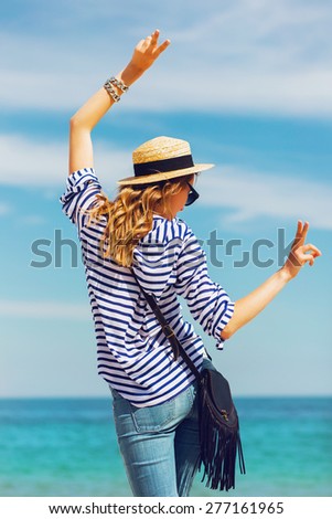 Young pretty blonde   woman standing on the beach near the sea back waiting and have fun. Wearing straw hat and striped shirt. Posing in sunny summer day.