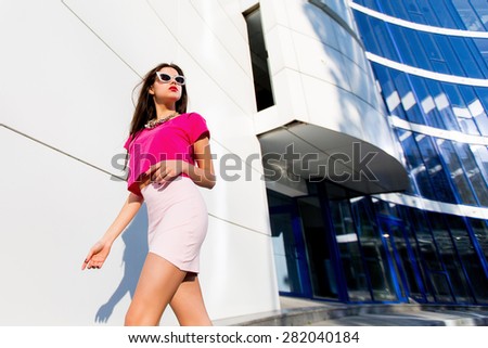 Fashion portrait of glamour bright sexy woman in pink top and skirt with perfect long legs walking against modern urban background.