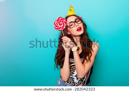 Crazy funny pretty woman posing against blue wall wearing trendy summer  outfit , cool cute glasses and party hat. Holding big pink lollypop .