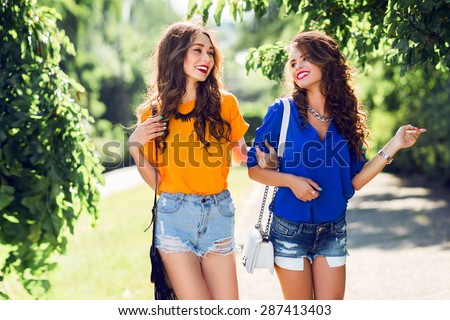 Two beautiful  girls walking in  the  summer park end talking.  Friends wearing stylish shirt and jeans shorts , enjoying  day off and have fun.