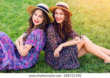 Outdoor  photo of two best friends   posing   on green meadow and  enjoy time together. Wearing straw hat and colorful boho dress. Summer sunny day.