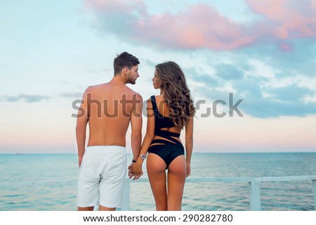 Young attractive  couple in love,  Handsome  man and sexy  woman with perfect tan slim fit  body  enjoying romantic evening on the beach, Holding hands watching  on the sea.