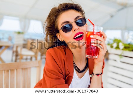 Close up portrait of cute pretty stylish young woman posing outdoor, sitting in summer cafe and drinking  exotic cocktail, sea background. Bright colors. Vacation mood. Smiling and have fun.