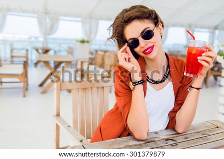 Close up lifestyle  portrait of cute stylish young woman posing outdoor, sitting in summer cafe and drinking  exotic cocktail, sea background. Bright colors. Vacation mood. Smiling and have fun.