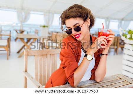 Close up lifestyle  portrait of cute pretty  young woman posing outdoor, sitting in summer cafe and drinking  exotic cocktail, sea background. Bright colors. Vacation mood. Smiling and have fun.