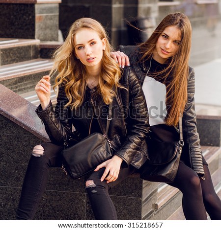 Two stylish pretty women posing on the street in sunny day. Wearing trendy urban outfit , leather jacket and boots heels. Young friends  waiting on stairs outdoor.