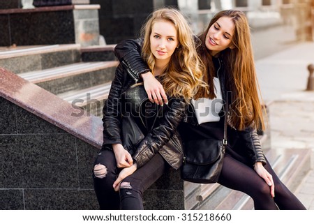 Fashion portrait of Two stylish pretty women posing on the street in sunny day. Wearing trendy urban outfit , leather jacket and boots heels. Young friends  waiting on stairs outdoor.