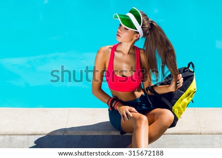 Fresh healthy young attractive woman in sport stylish outfit sitting near pool on summer hot day . Have  perfect tan slim fit body . Bright neon  backpack.
