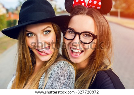 Close up lifestyle  portrait  of  girls best friends makes funny  grimaces on camera , show tongue  and laughing together.Two women posing outdoor, warm sunny evening colors.