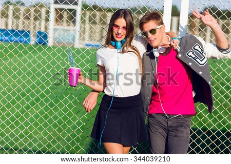 Young  friends funny guys active people have fun together, girl and guy summer urban casual style. Slim tan pretty woman with bright make up, stylish headphones  near sports ground.