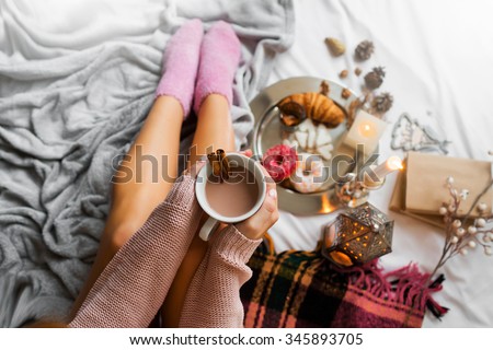 Woman relaxing at cozy home atmosphere on the bed. Young woman with cup of coffee or cocoa in hands and cookies enjoying comfort. Soft light and comfy lifestyle concept.