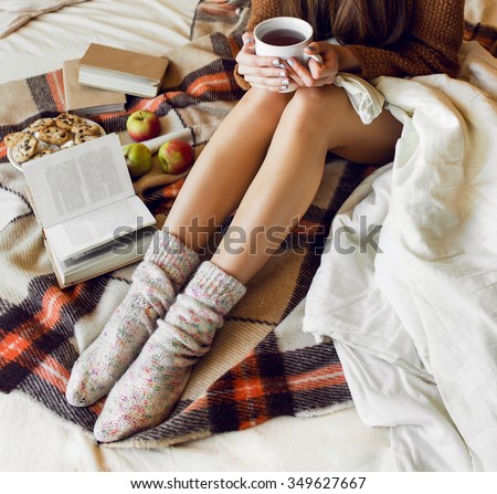 Soft cozy photo of slim tan woman in warm sweater and woolen socks  on the bed with  book  in hands, top view point.  Checkered plaid near  a plate of oatmeal cookies. Fall or winter time  concept.