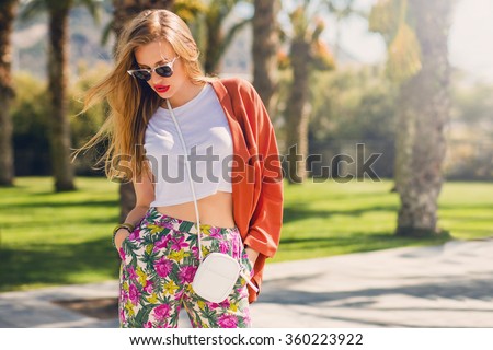 Amazing glamour blonde woman in trendy summer outfit posing outdoor palm trees background. Bright make up, orange jacket, sunglasses, bag in hands. Fashionable model walking in park .