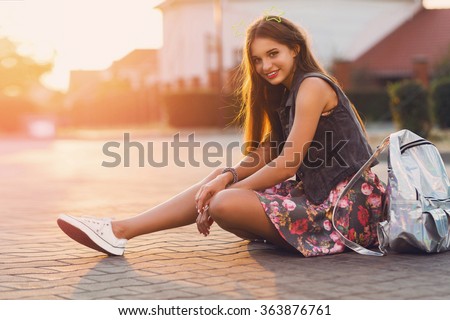 Young fresh cheerful hipster  girl  sitting on the road in countryside wearing stylish summer clothes, bright print skirt , neon  bag pack. Warm evening colors. Sunset. Lifestyle image.