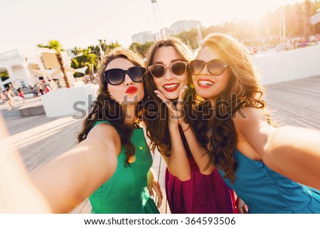 Lifestyle sunny image of best friend girls  taking selfie on  camera, crazy emotions , happy vacations, shopping  day. Wearing elegant dress, way hairstyle, sunglasses and red lips.