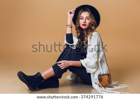 High fashion portrait of young elegant blonde  woman in black wool hat  wearing oversize white fringe  poncho with long grey dress. Studio shot. American hippie bohemian style.