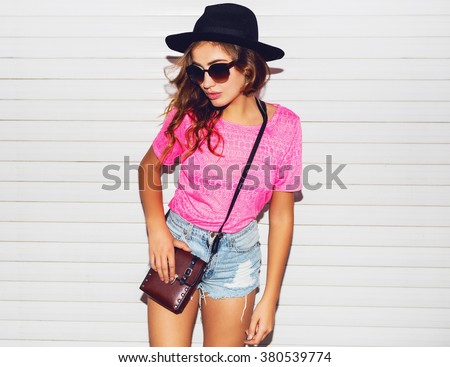 Stylish night flash fashion portrait of trendy  casual young  woman in pink neon  t-shirt , black hat, stylish shirts posing near white urban  wall along .