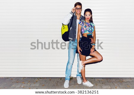 Fashion image of  stylish young  couple in love , tourists , posing near white urban wall. Wearing cool spring clothes, vintage camera,  bright neon back pack , sneakers.