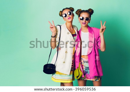 Fashion studio image of two young women in stylish casual  spring outfit   having fun, show  tongue. Bright trendy pastel  colors, stylish hairstyle  with buns , cool sunglasses. Friends portrait.