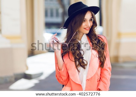 Outdoor  fashion portrait of stylish  pretty brunette  woman in night spring casual outfit walking in the city. Wearing white blouse, pink jacket, black wool hat. Long wavy hairstyle.