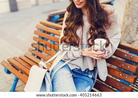 Outdoor fashion details. Sunny lifestyle  image of   stylish young  attractive woman in cozy knitted sweater  , holding cup of  hot  beverage , sitting on wooden  city bench.