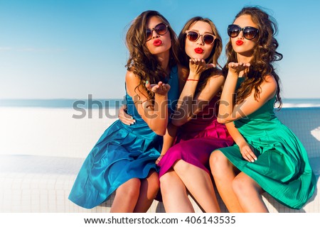 Three  beautiful   glamour  girls  in same colorful dresses posing near the beach, wearing stylish sunglasses and enjoying vacation in summer city. Bright colors. Casual outfit. Send kiss.