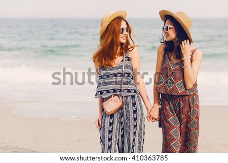 Portrait of  two young female friends walking on the sea shore looking at camera laughing, talking. Sisters  strolling along a beach. Stylish summer beachwear, straw hat , sunny colors.